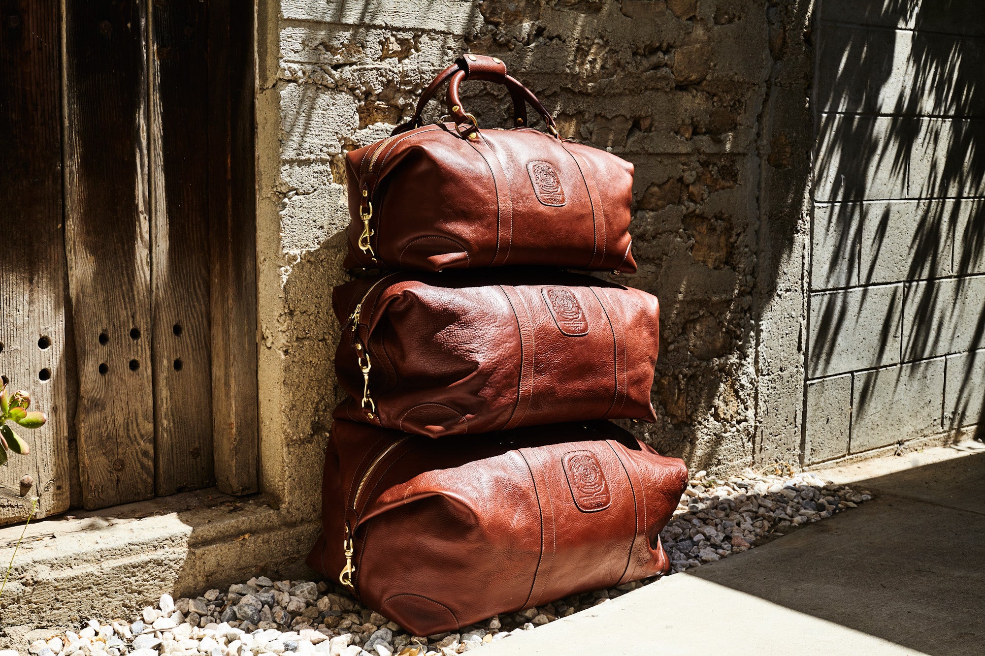 Three leather duffle bags stacked in front of a stone building with a wooden door.