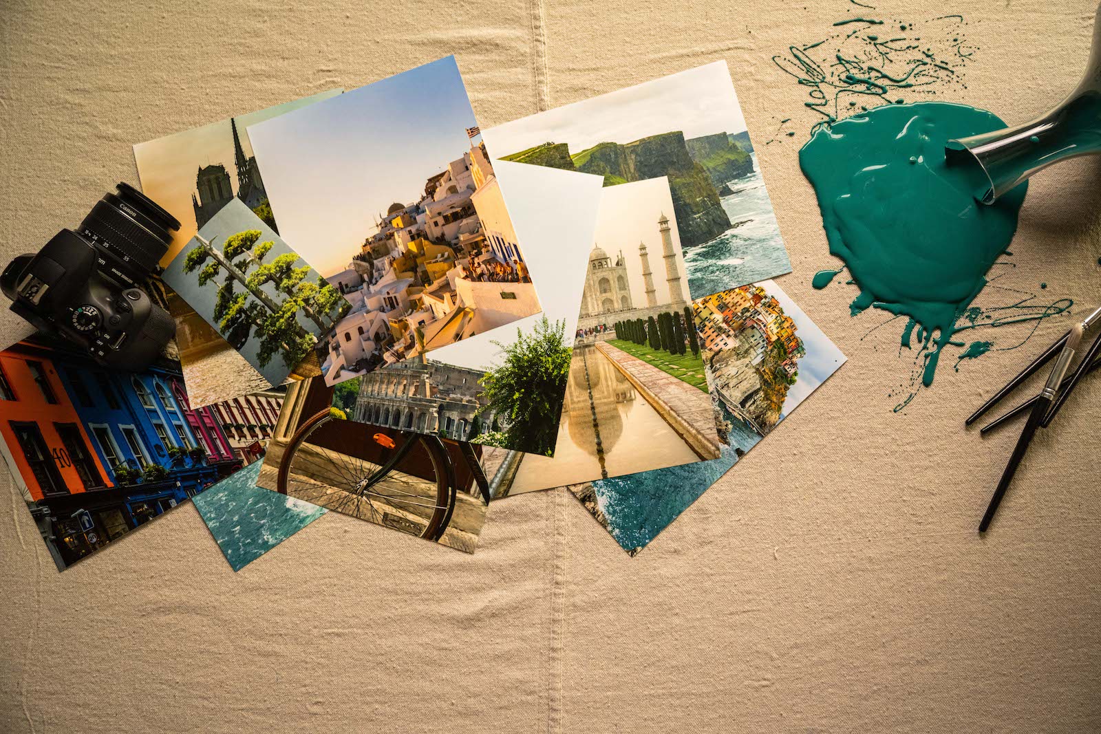 a pile of travel photos sitting on beige tablecloth with bottle of spilled green paint and paint brushes